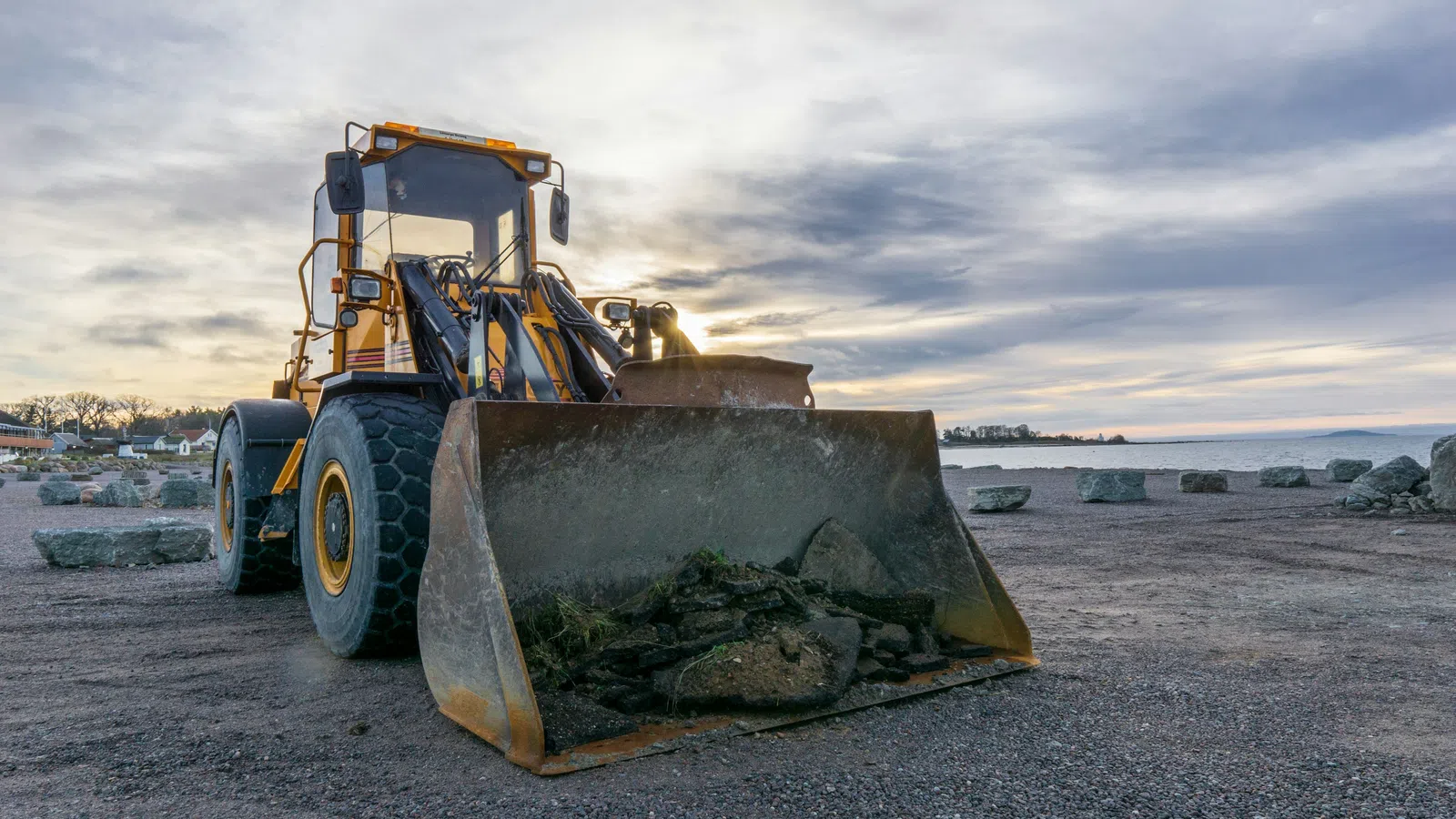 Heavy yellow front loader with dirt and rocks in bucket at a construction site during sunset.