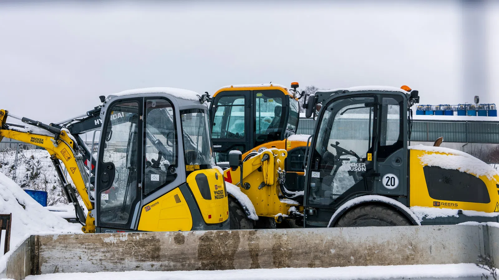 Three yellow and black construction vehicles covered with snow parked side by side in a snowy lot.