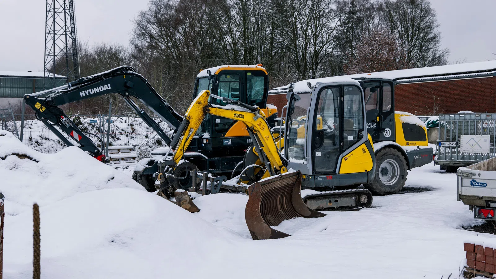 Yellow and black excavators parked on snow-covered ground near construction materials and bare trees in winter.