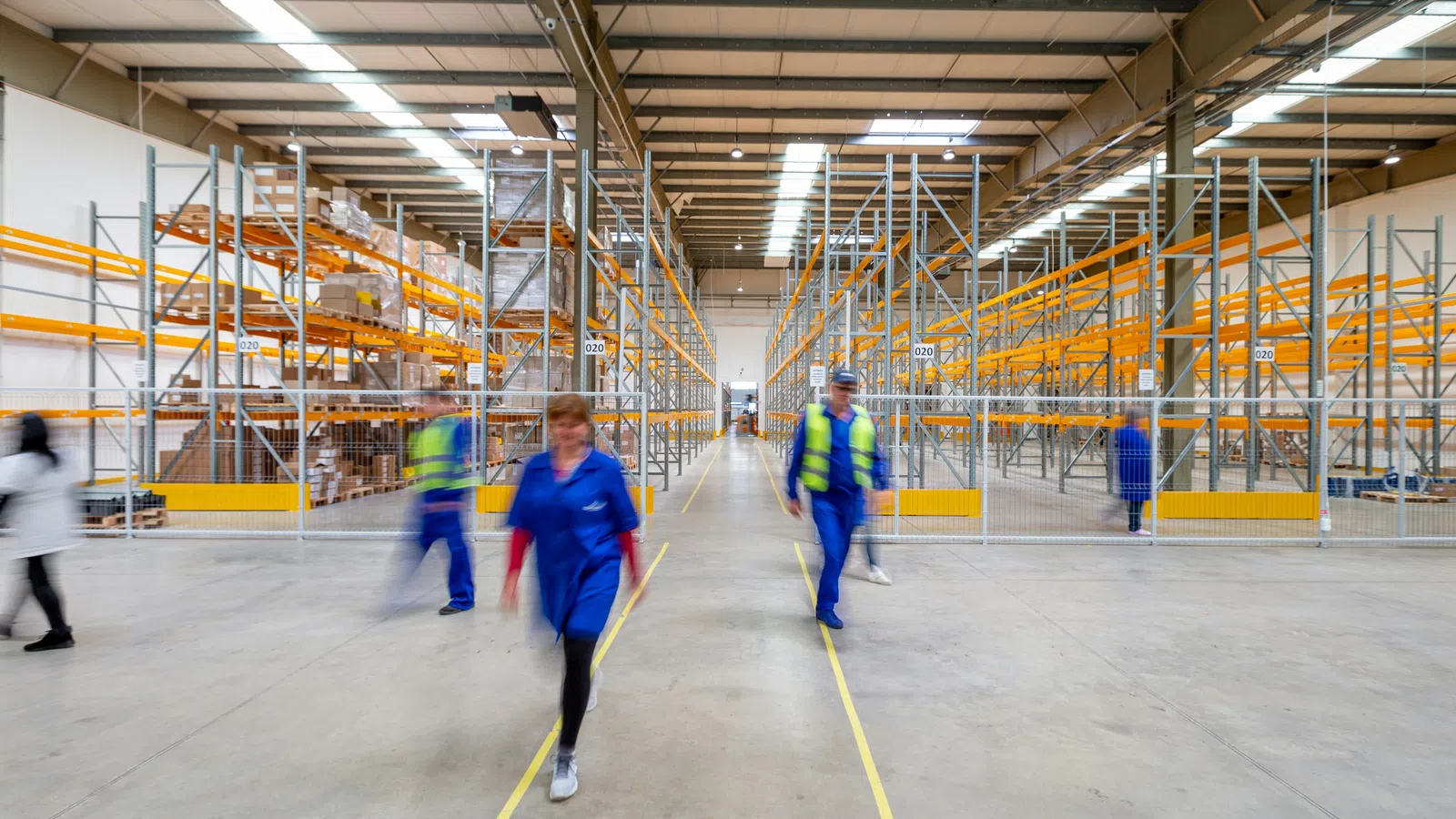 Workers moving inside a spacious warehouse with tall orange shelving and high ceilings.