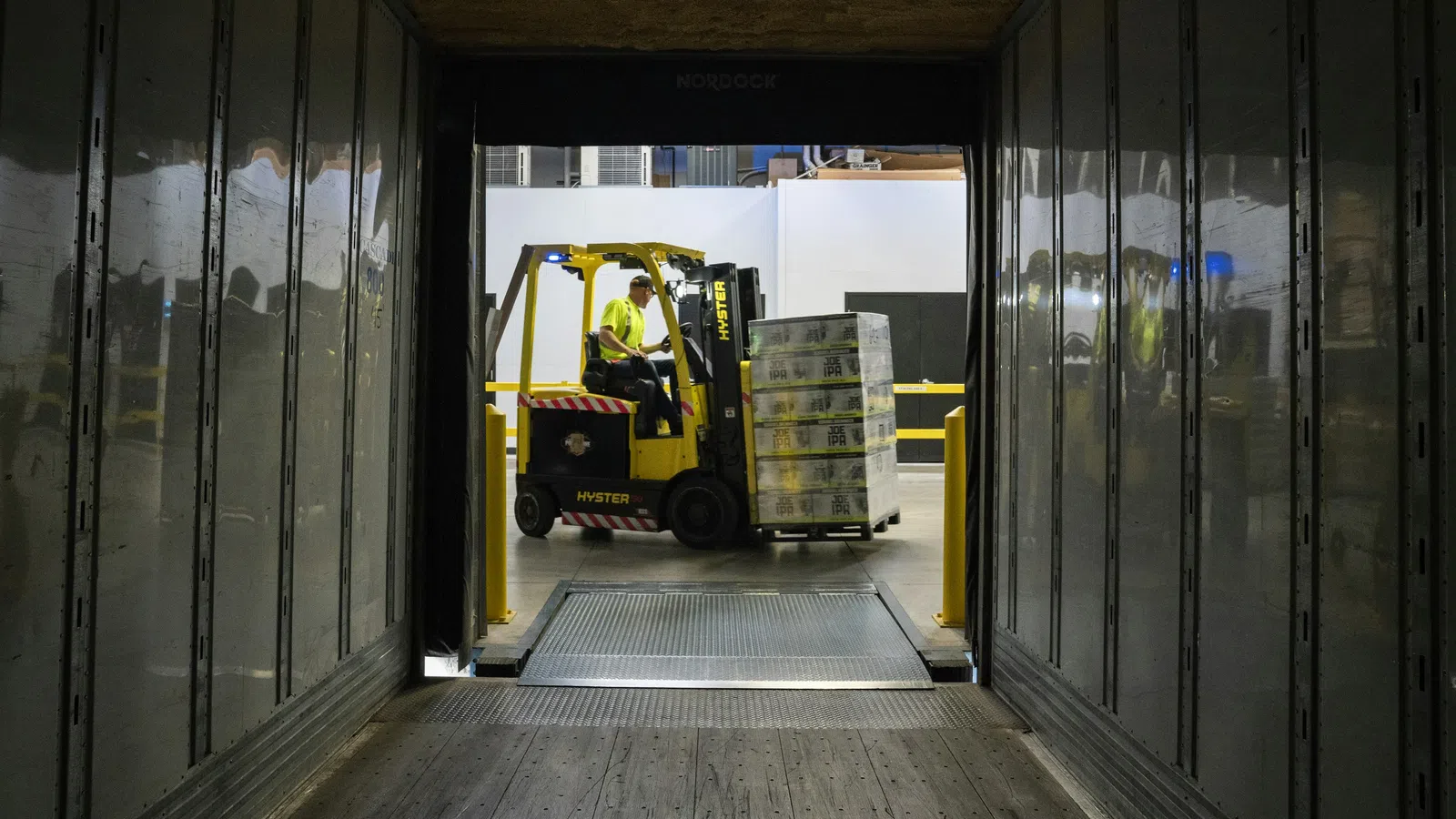 Forklift operator loading stacked pallets into a trailer through a warehouse loading dock with metal walls.