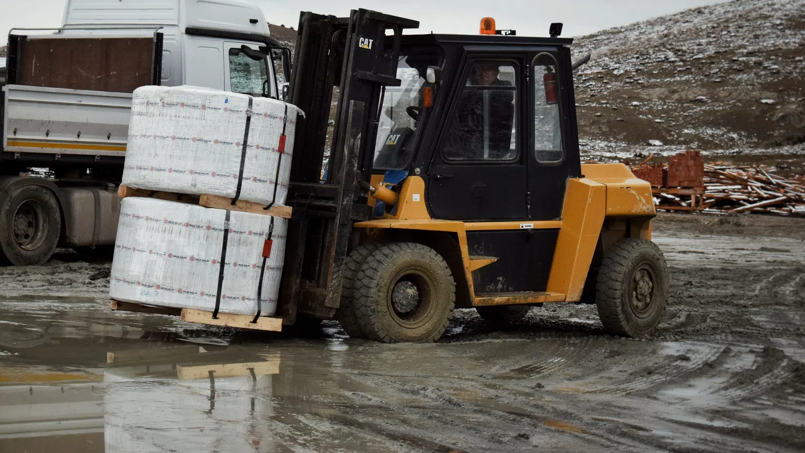 Yellow forklift lifting wrapped pallets in an outdoor muddy industrial area with a white truck in the background.