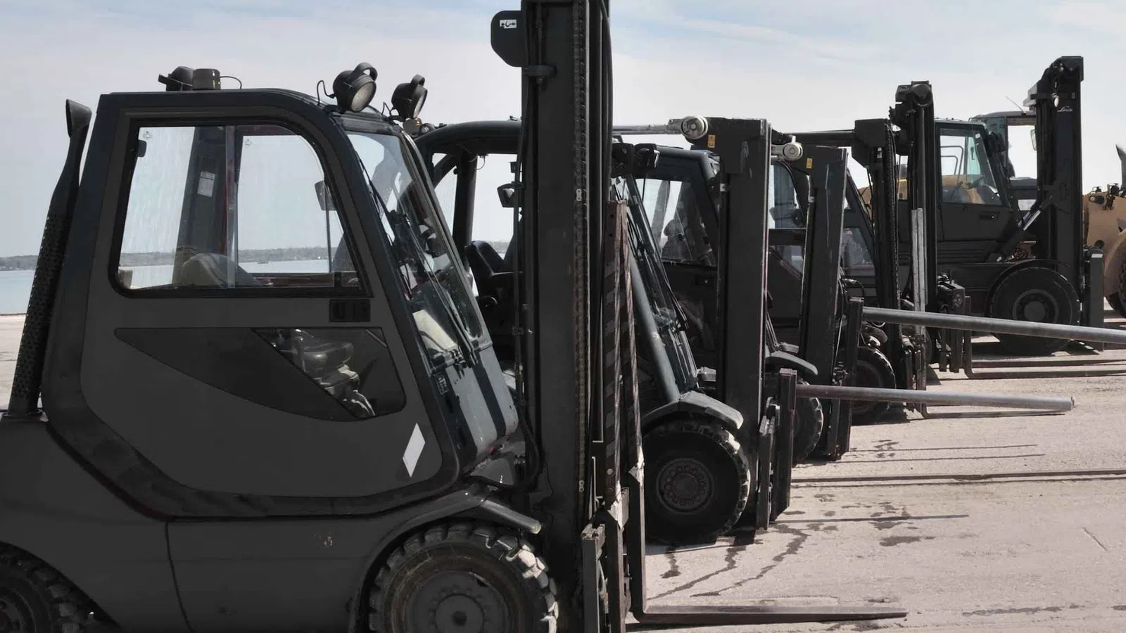 Row of black forklifts parked in a line outdoors on concrete under a cloudy sky.