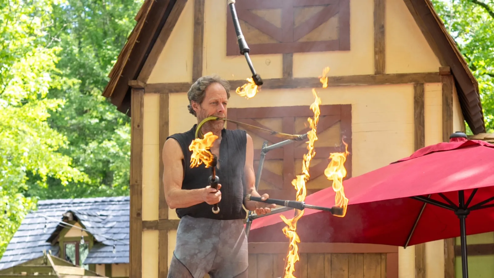 Male fire juggler balancing on a rope over flames in a rustic outdoor setting with wooden structures.