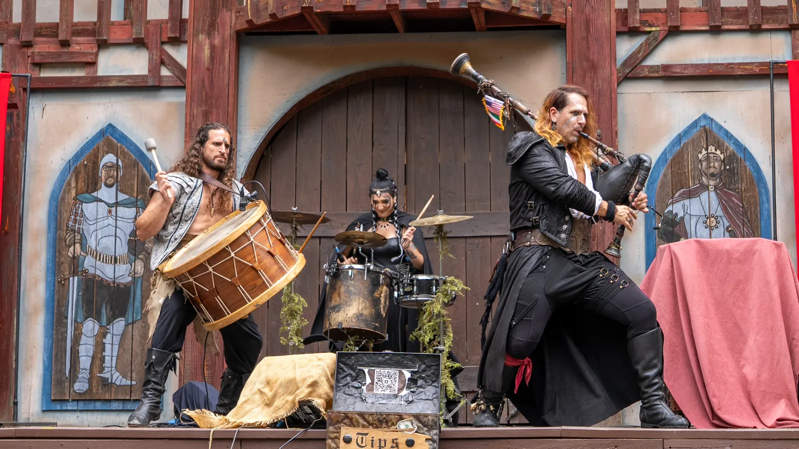 Medieval band performing with bagpipes, drums, and violin on rustic outdoor stage with knight motifs and red banners.