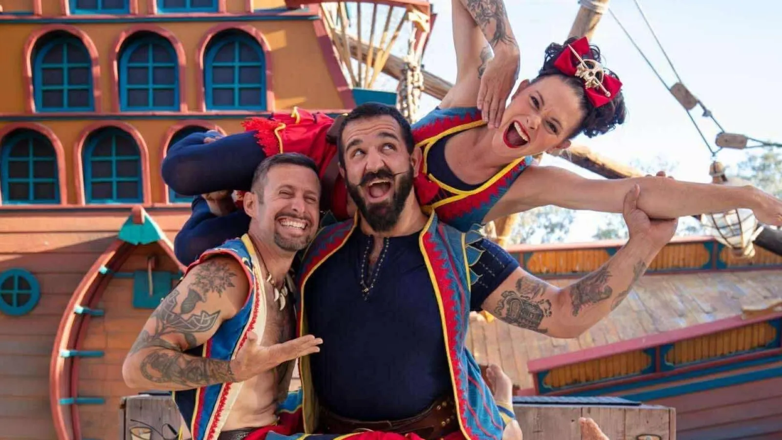 Three joyful performers in colorful pirate costumes posing playfully on a ship-themed stage outdoors.