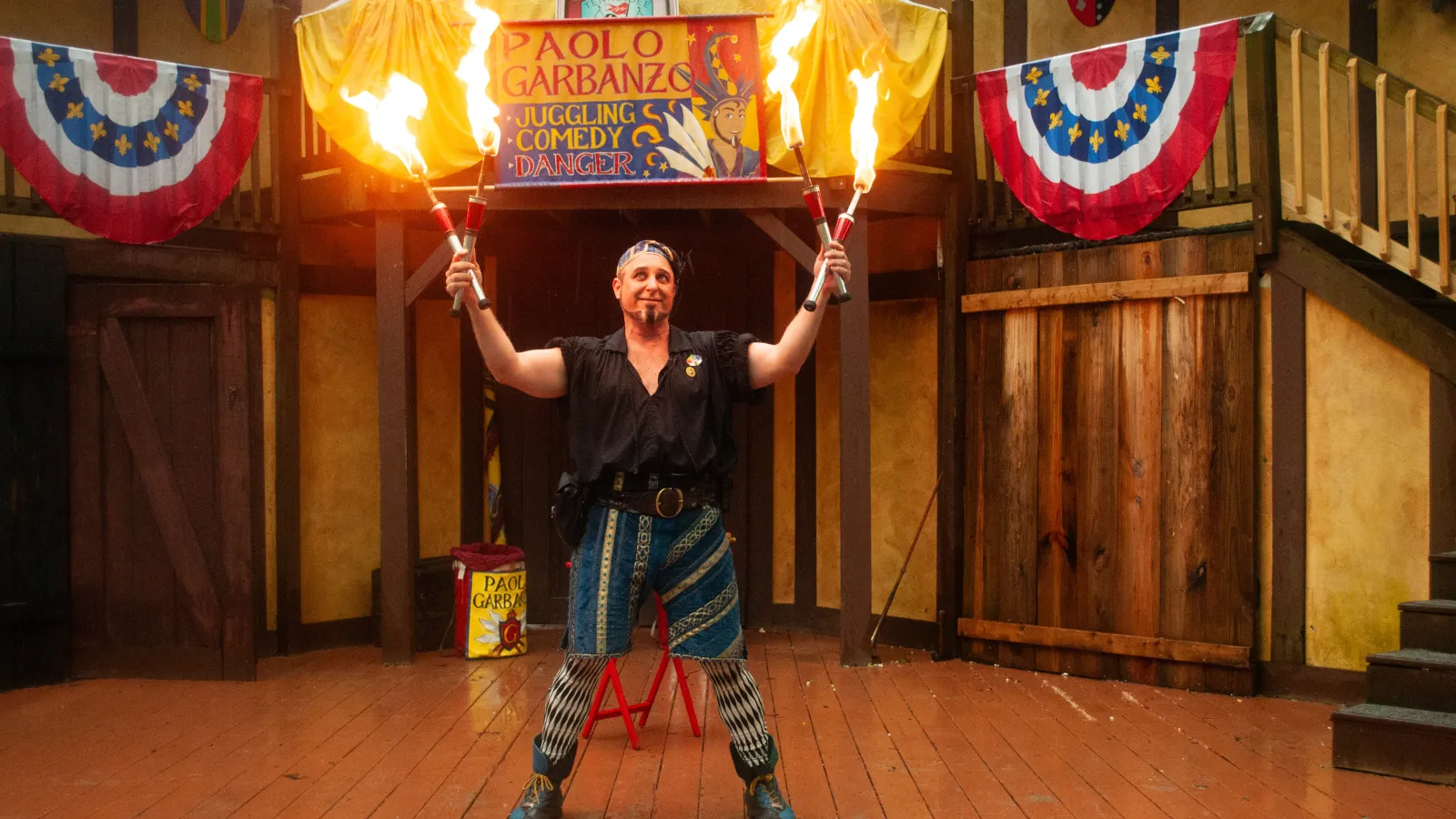Fire juggler holding four flaming torches on stage with colorful banners and wooden background.