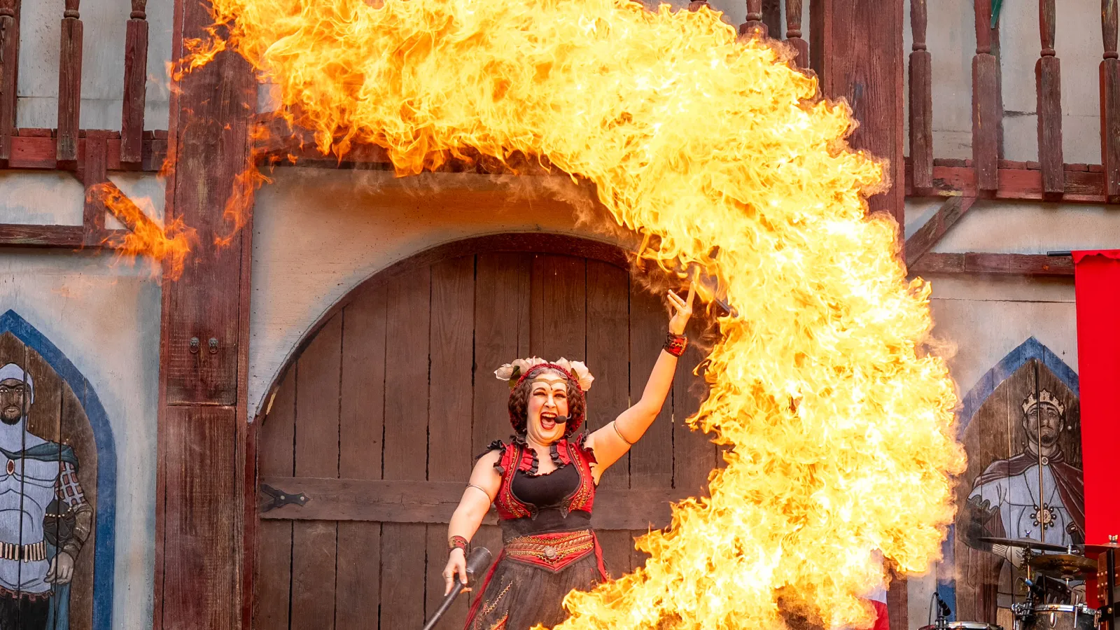 Female fire performer creating a large flame arc on stage at a medieval-themed event with wooden backdrop