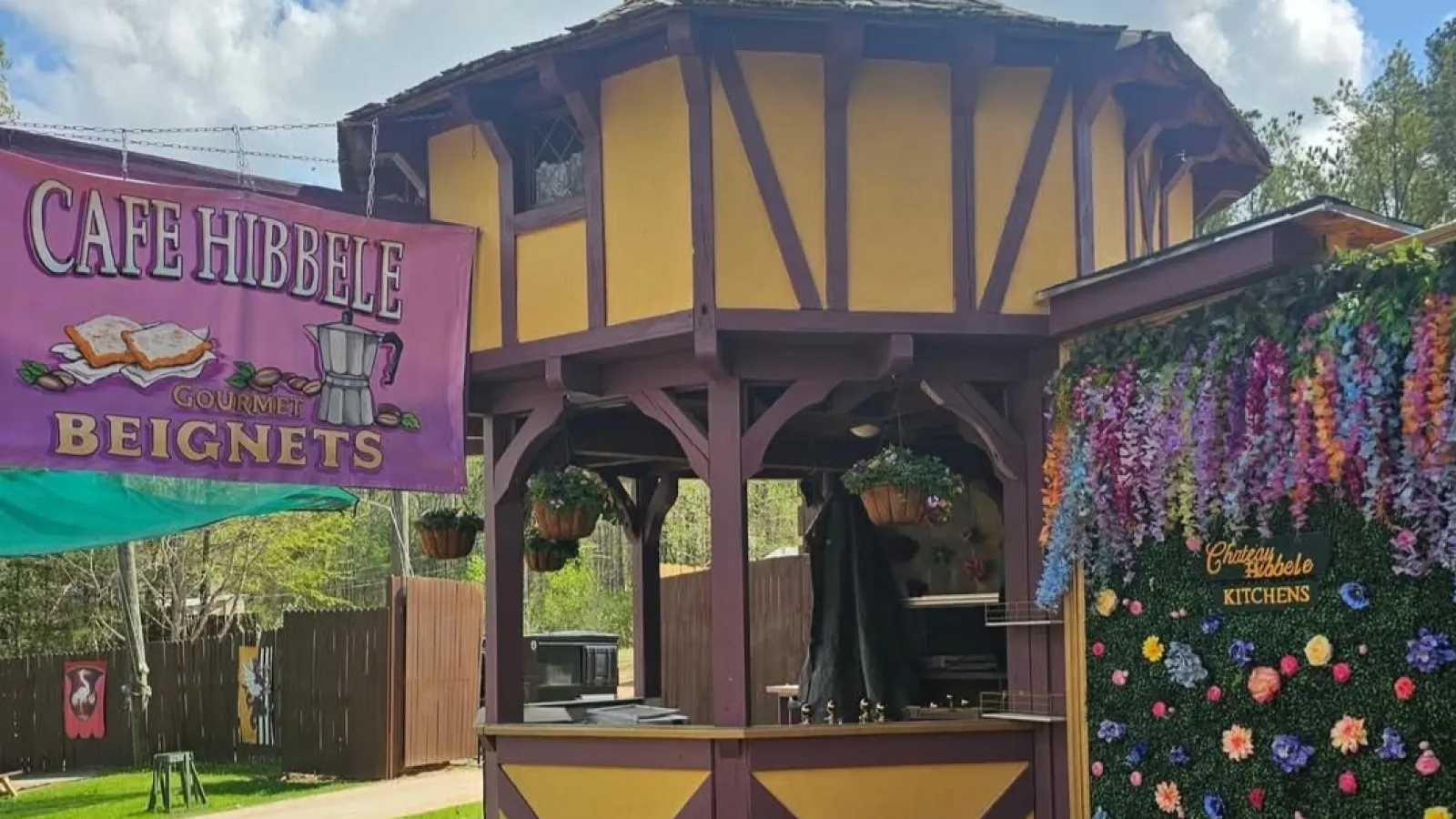 Yellow and brown wooden gazebo at Cafe Hibbele with hanging plants and flower wall under blue sky.