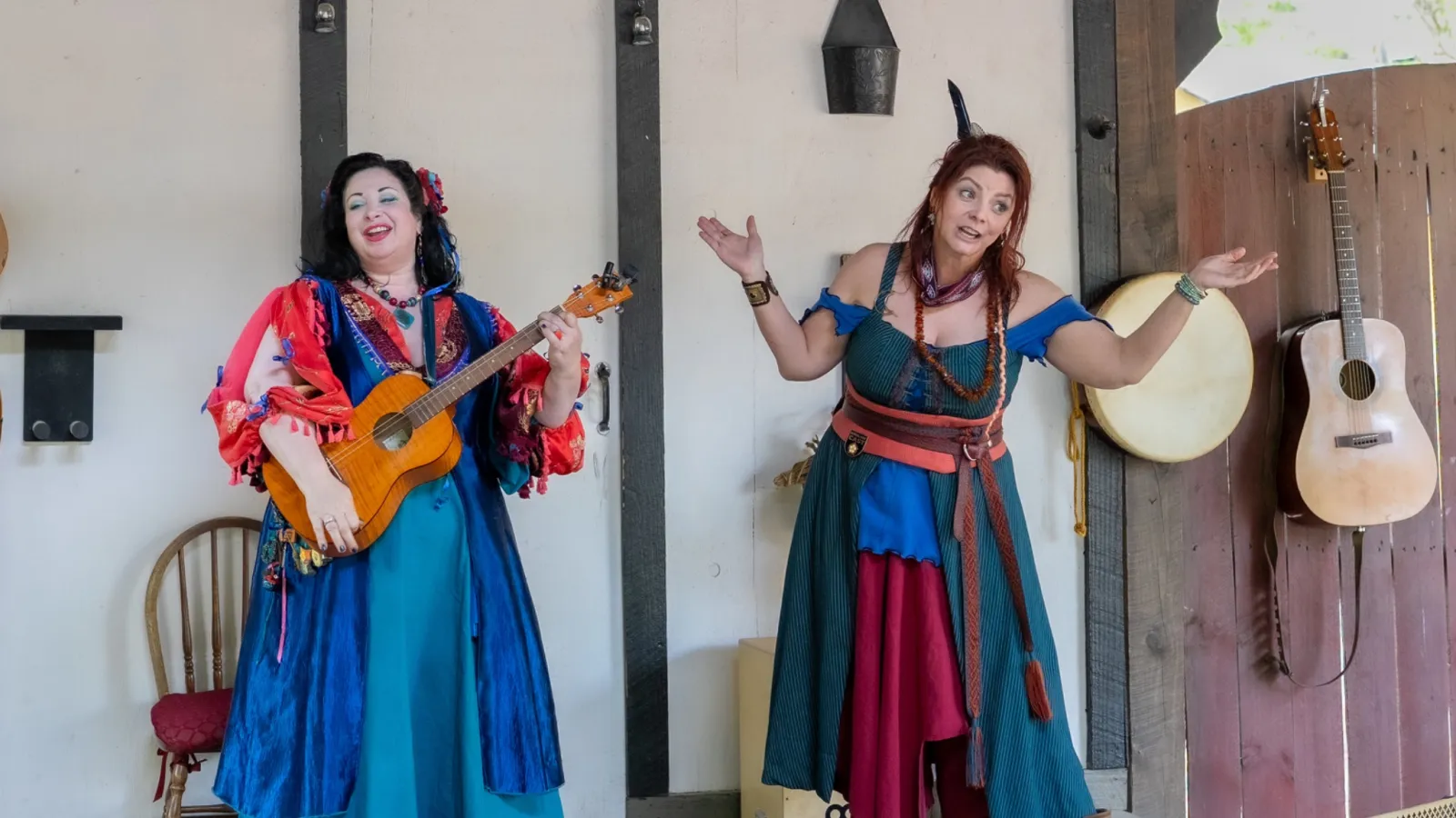 Two women in colorful Renaissance costumes perform music and storytelling on a rustic indoor stage.