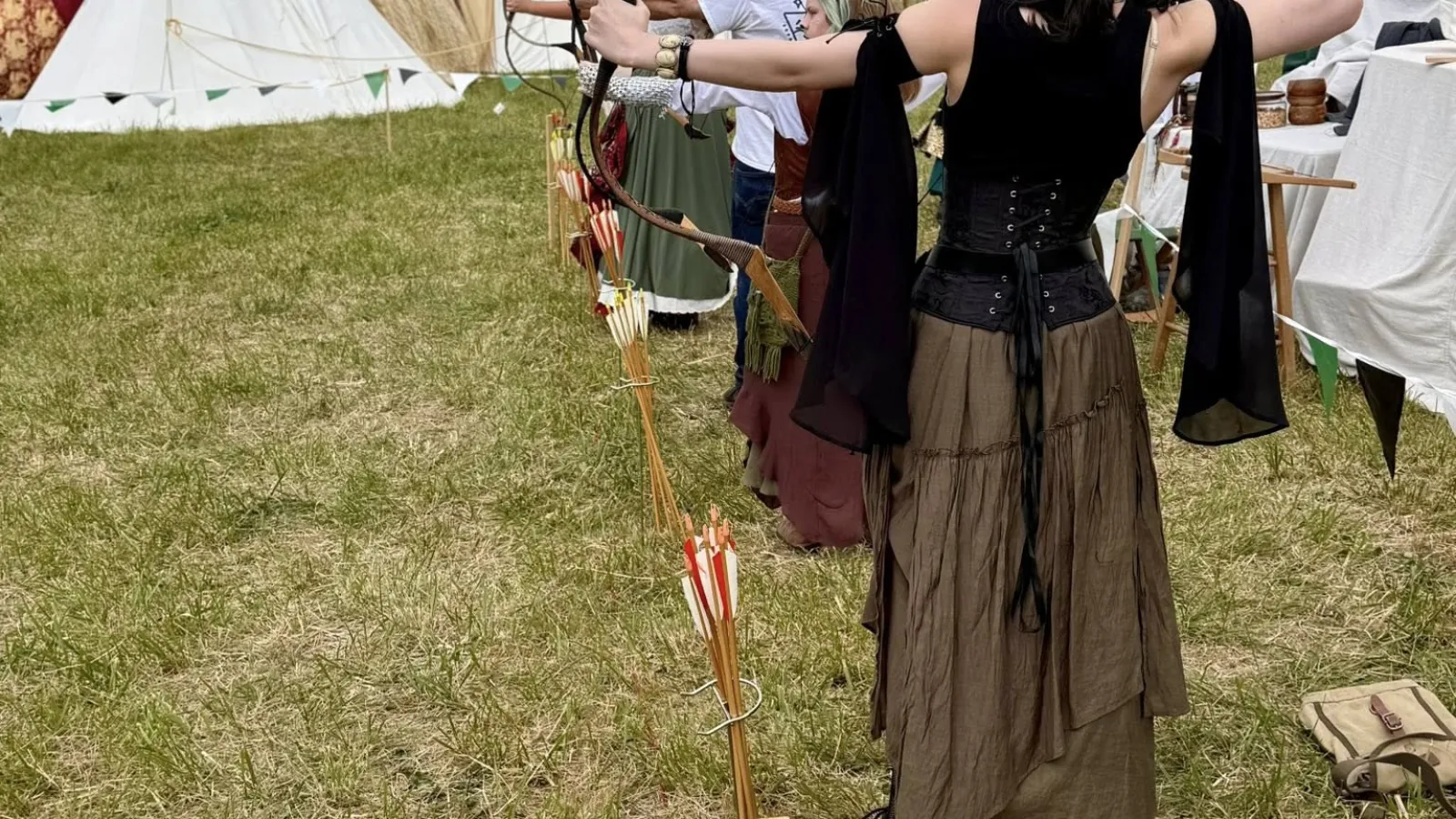 Group of archers in medieval-style clothing aiming bows at an outdoor archery event with tents and bunting in the background