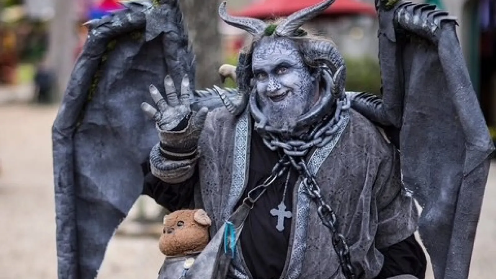 Person in detailed horned and winged fantasy costume with gray makeup and teddy bear on shoulder at outdoor event.
