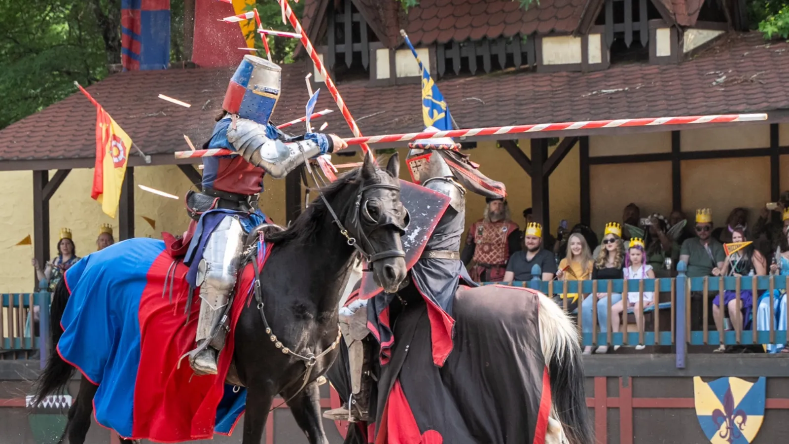 Two knights in armor jousting on horses with spectators watching at a medieval festival event.