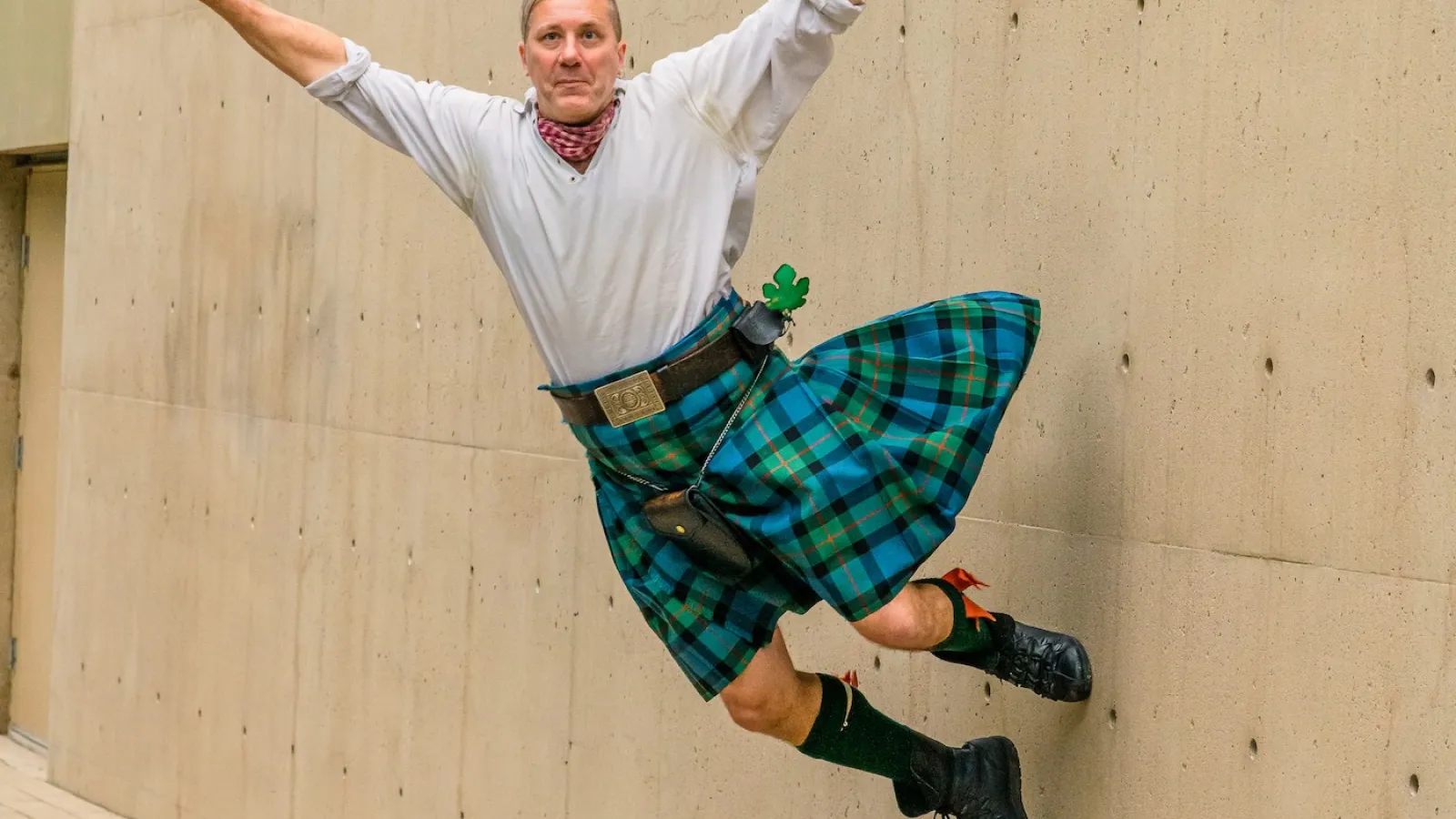 Man in a blue plaid kilt jumping mid-air against a concrete wall on