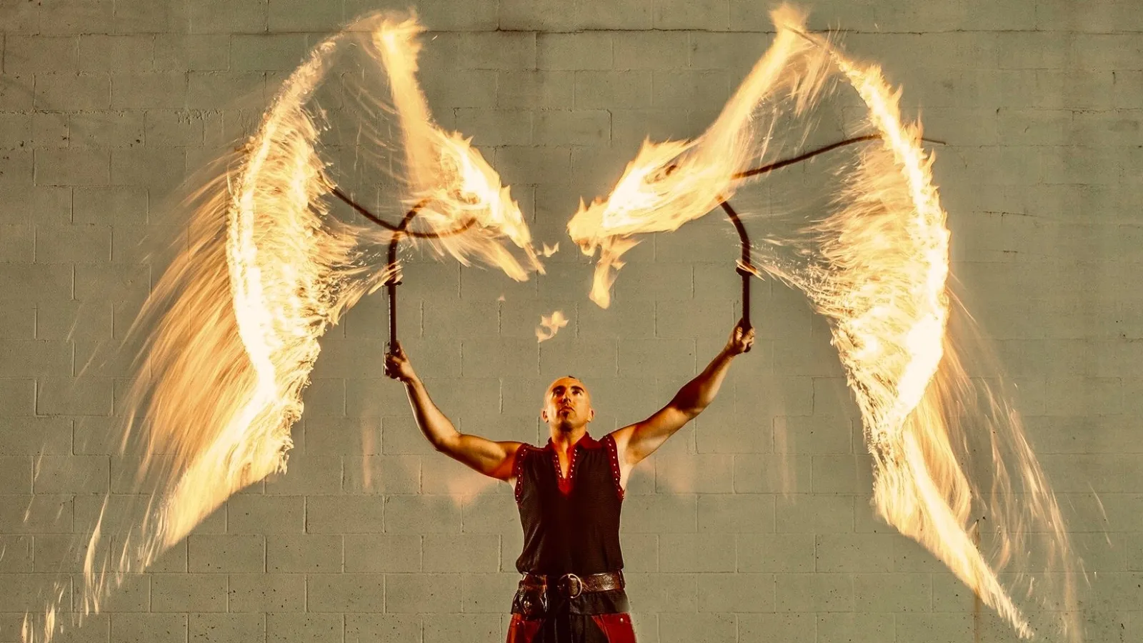 Fire performer creating arches of flames with two fire staffs against a plain wall background.
