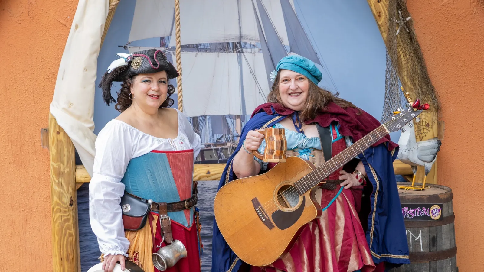 Two women in colorful pirate costumes holding a tambourine and guitar with a ship backdrop at a festival.