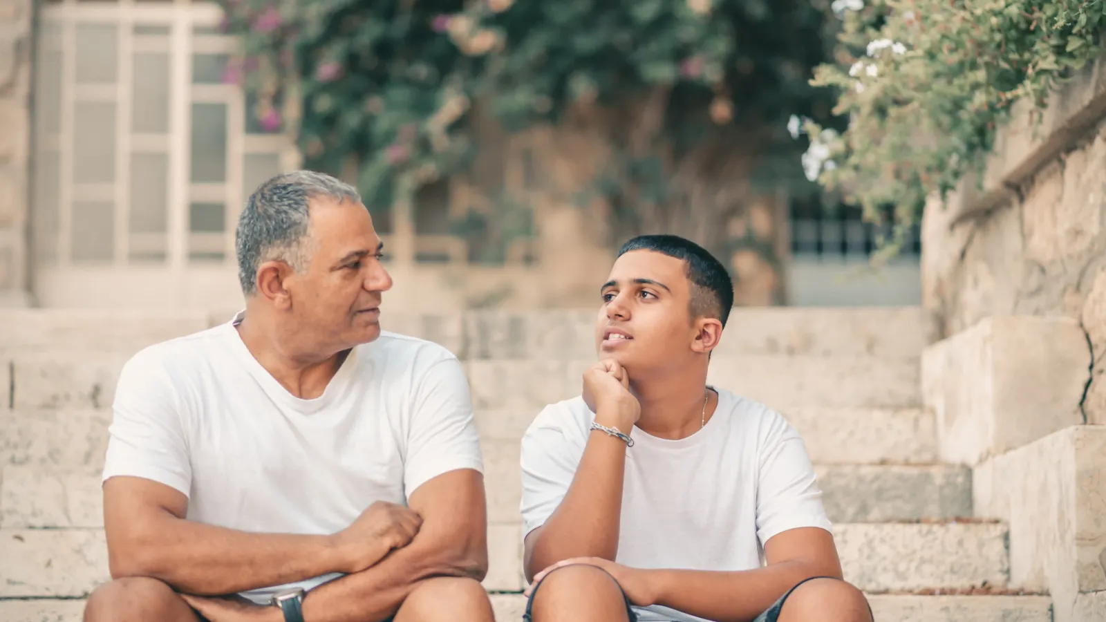 Adult man and teenage boy wearing white t-shirts sitting on stone steps having a conversation outdoors.