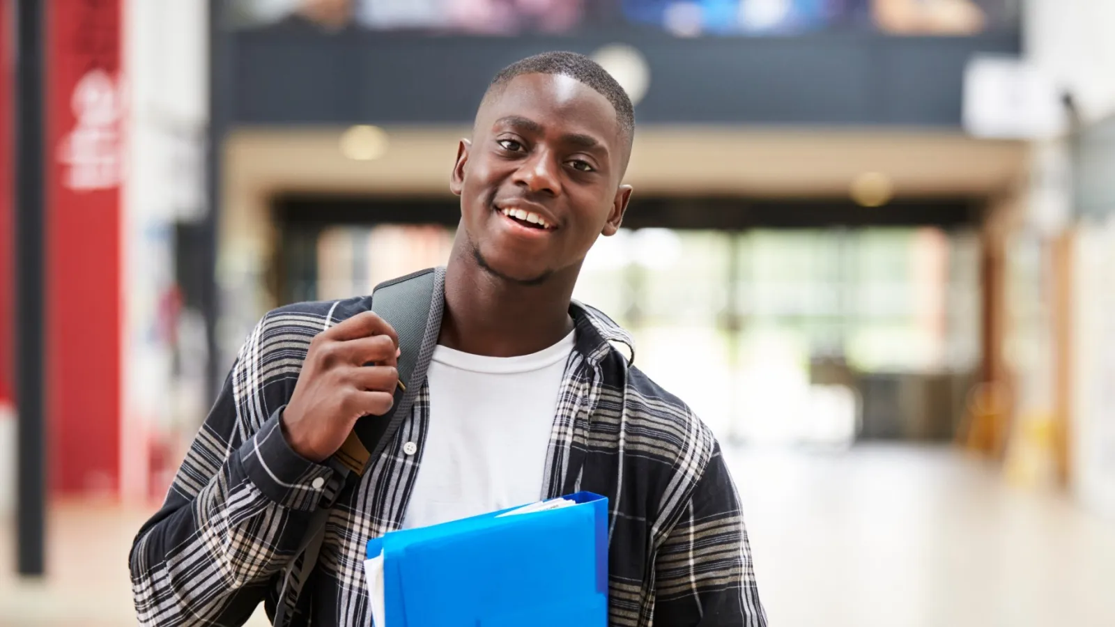 Smiling male student carrying blue folder and backpack walking through bright, modern school hallway.