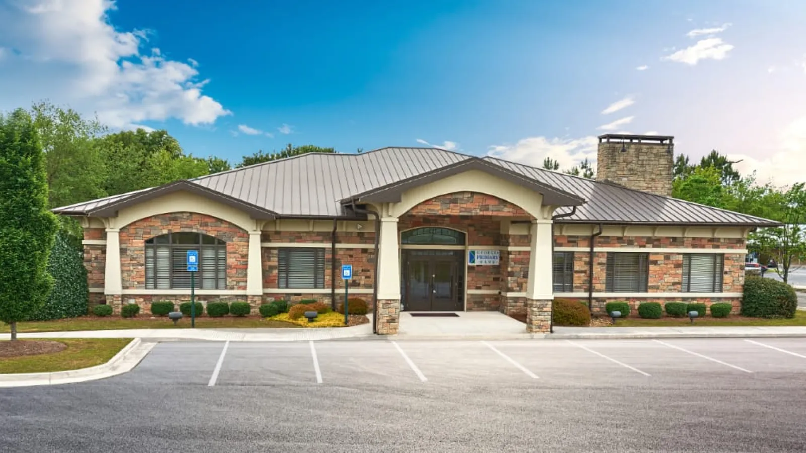 Modern brick building with a gray metal roof and landscaped parking area under a sunny sky.