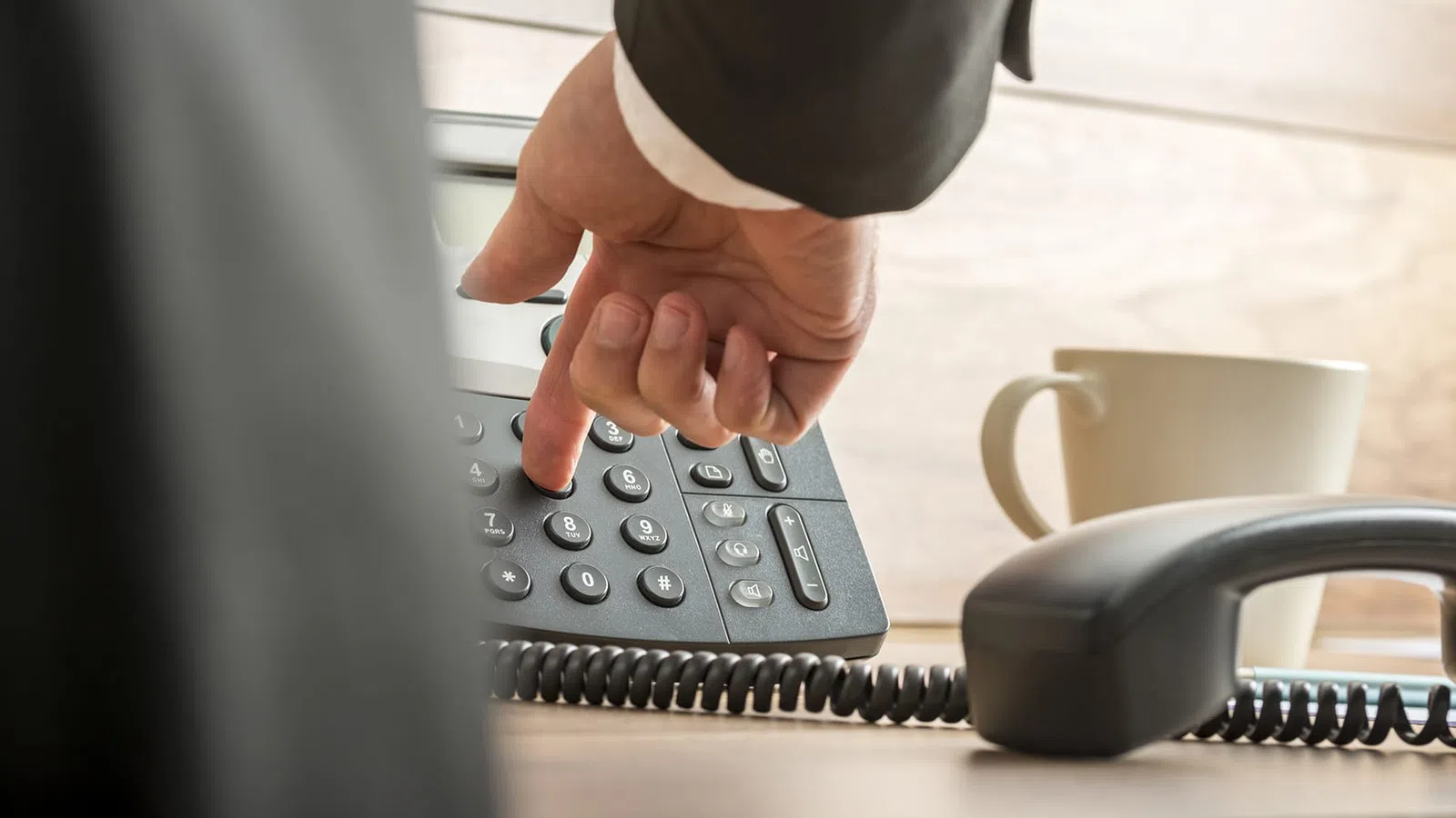 Person dialing a number on a desk phone with a coffee cup in the background in an office setting