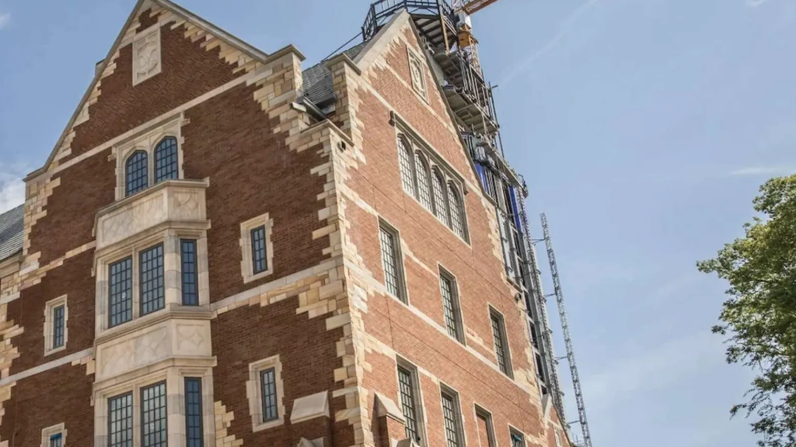 Historic brick building under construction with cranes and scaffolding against a partly cloudy sky.