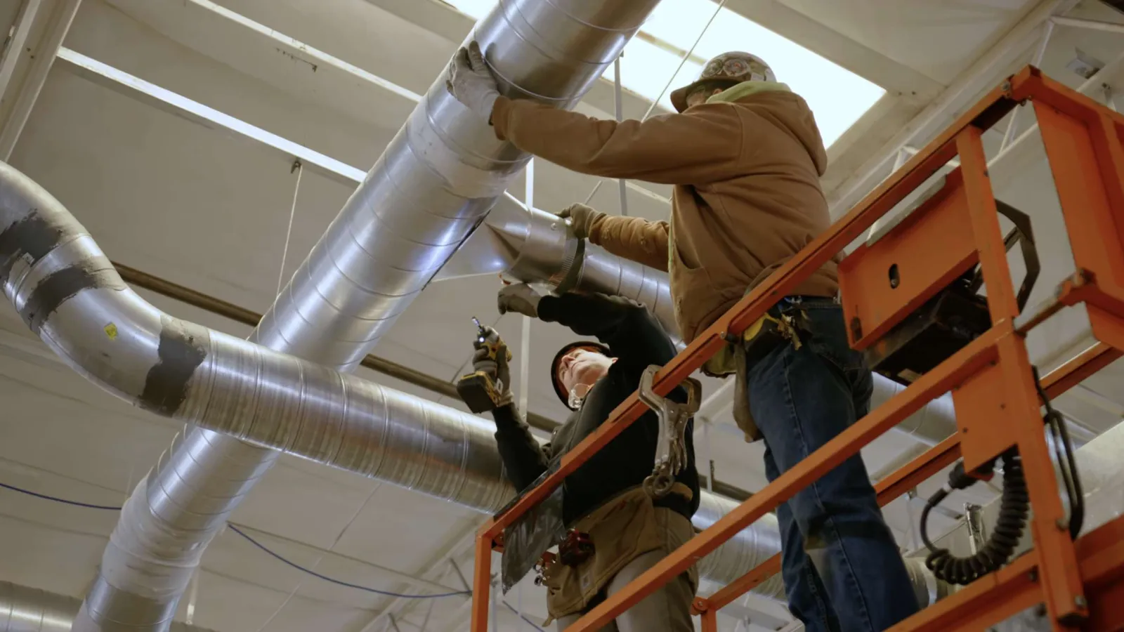 Two workers install ductwork inside a building using a scissor lift and power tools for HVAC setup.