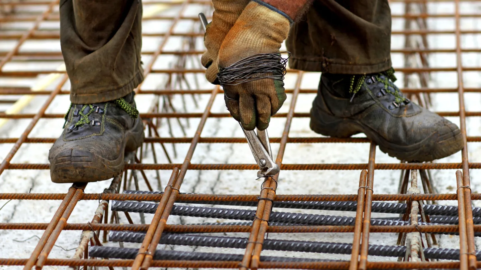 Construction worker fastening steel rebar with pliers on concrete foundation at building site.