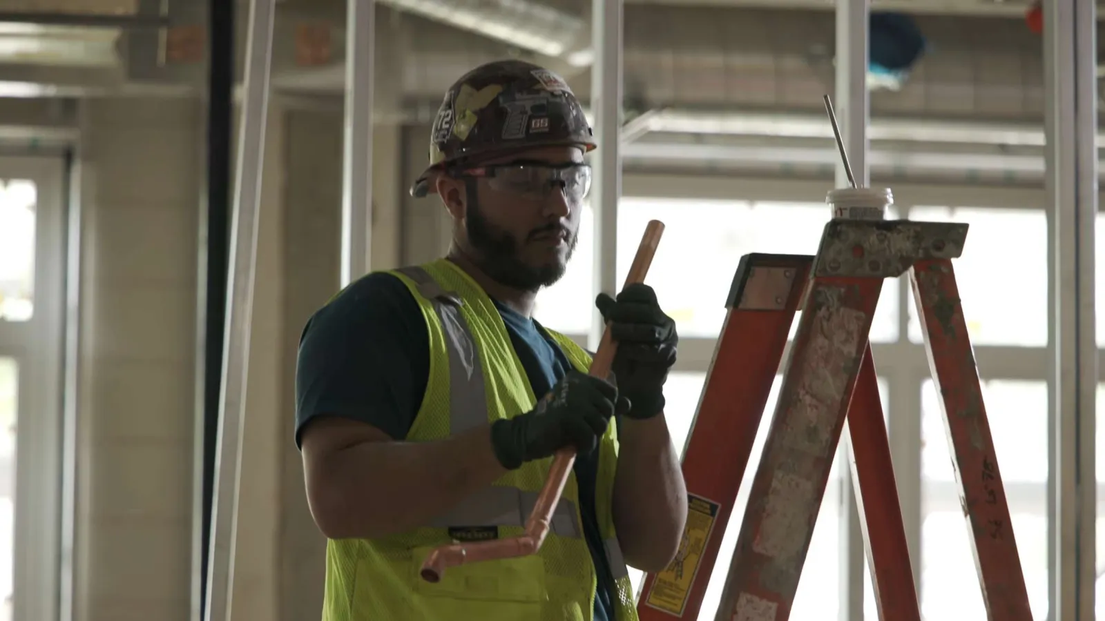 Construction worker in safety gear holding a pipe near a ladder inside a building under construction.