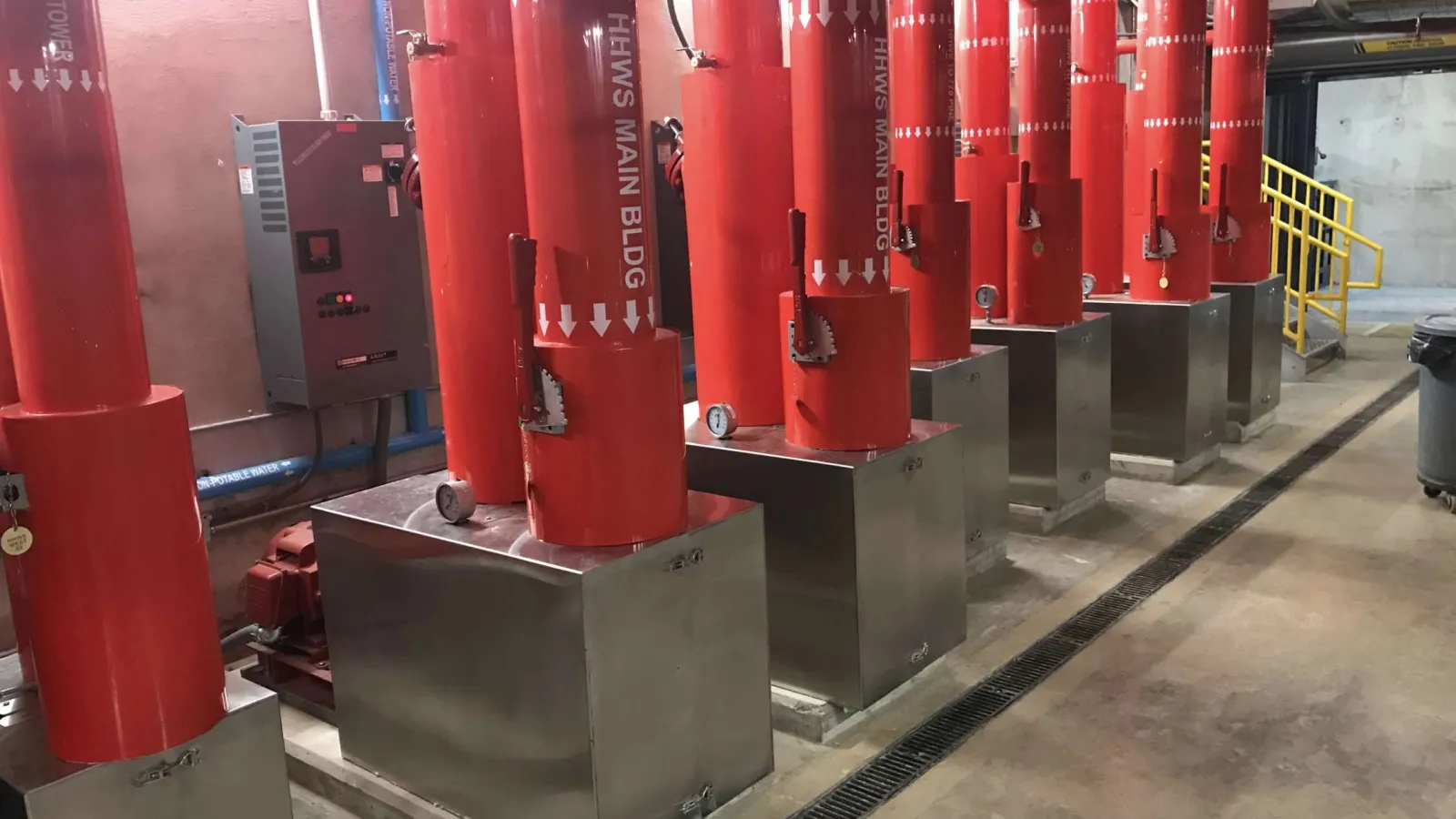 Row of large red pipes connected to metal bases in an industrial building with concrete floor and yellow stairs.