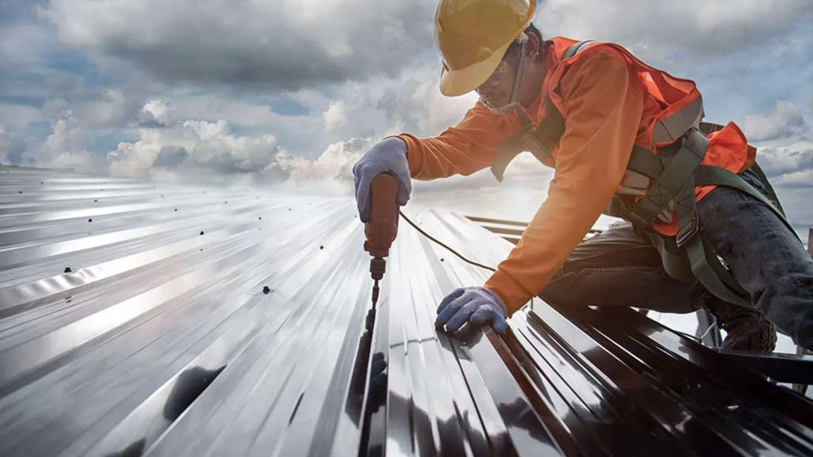 Construction worker in safety gear using power tool to install metal roofing under a cloudy sky outdoors.