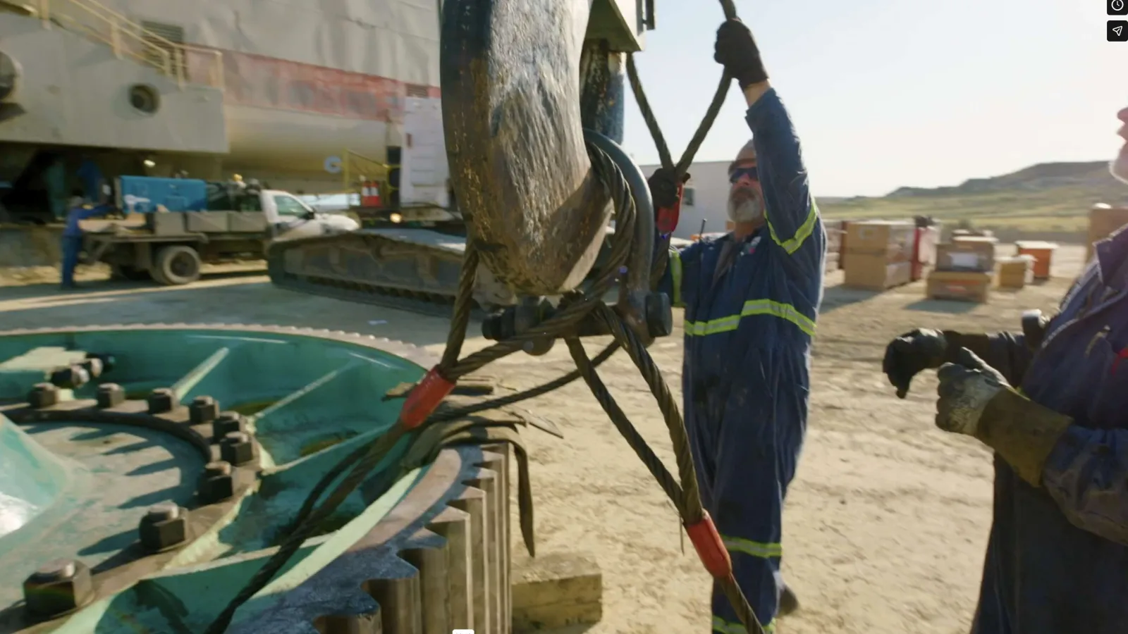 Workers in safety gear handling heavy lifting equipment on industrial site with large gear and machinery parts.