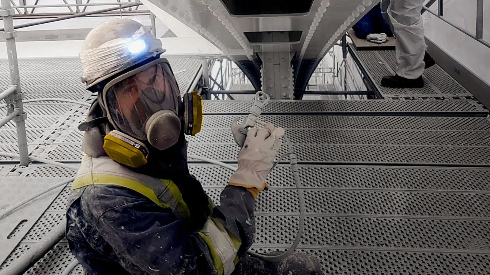 Worker in protective gear spray painting the underside of a metal bridge structure on an industrial platform.
