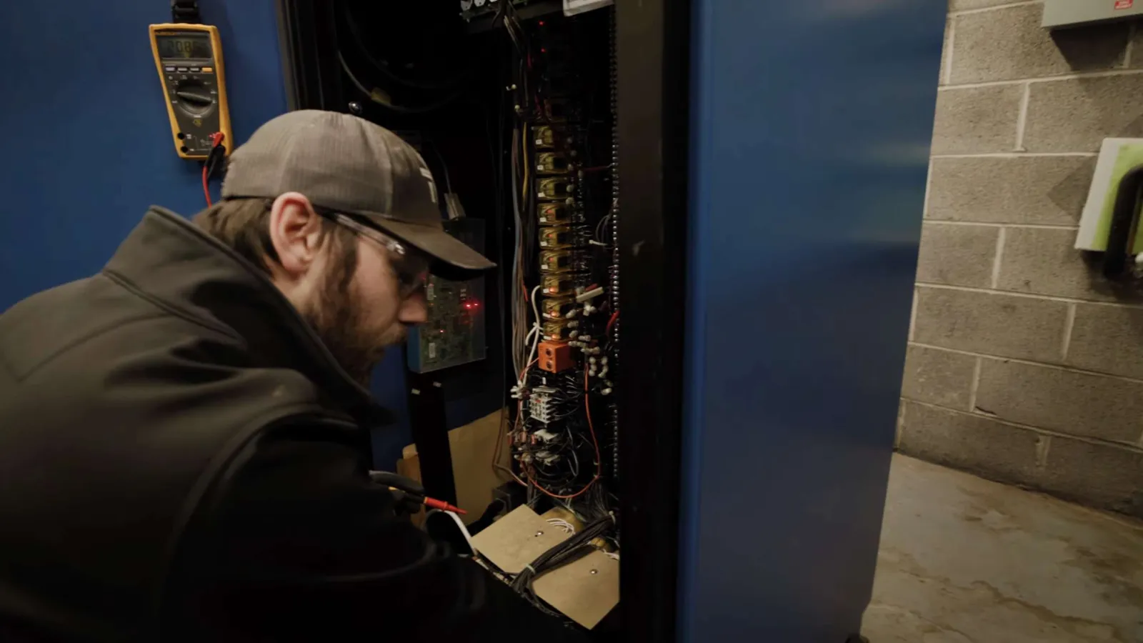 Technician wearing cap and glasses working on an open electrical control panel with wiring and equipment.
