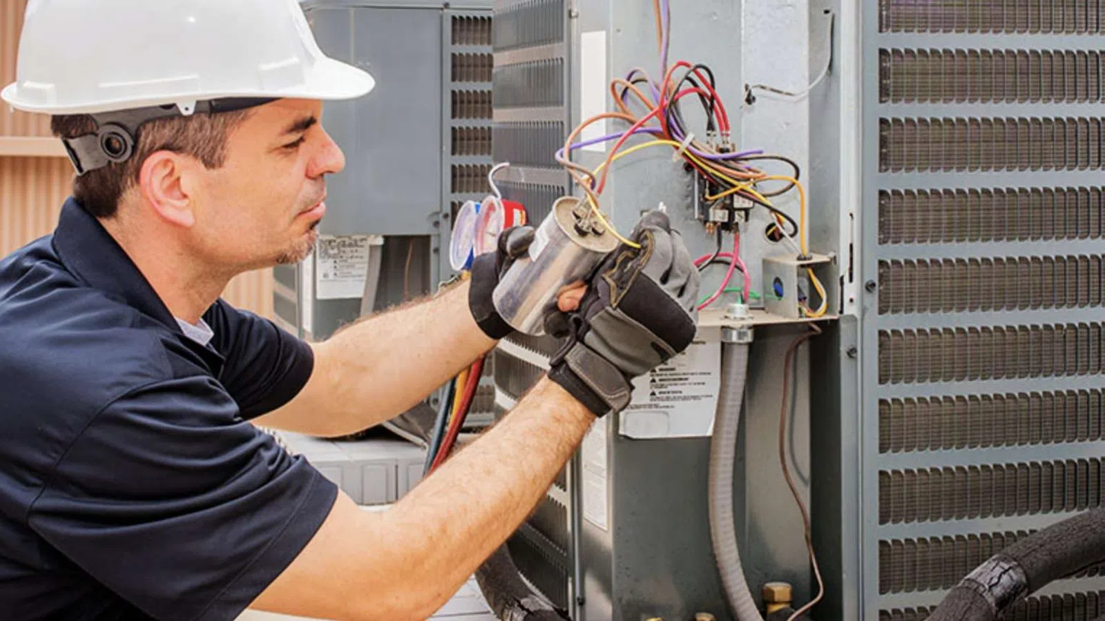 Technician in hard hat and gloves working on an outdoor HVAC unit with exposed wiring and components.