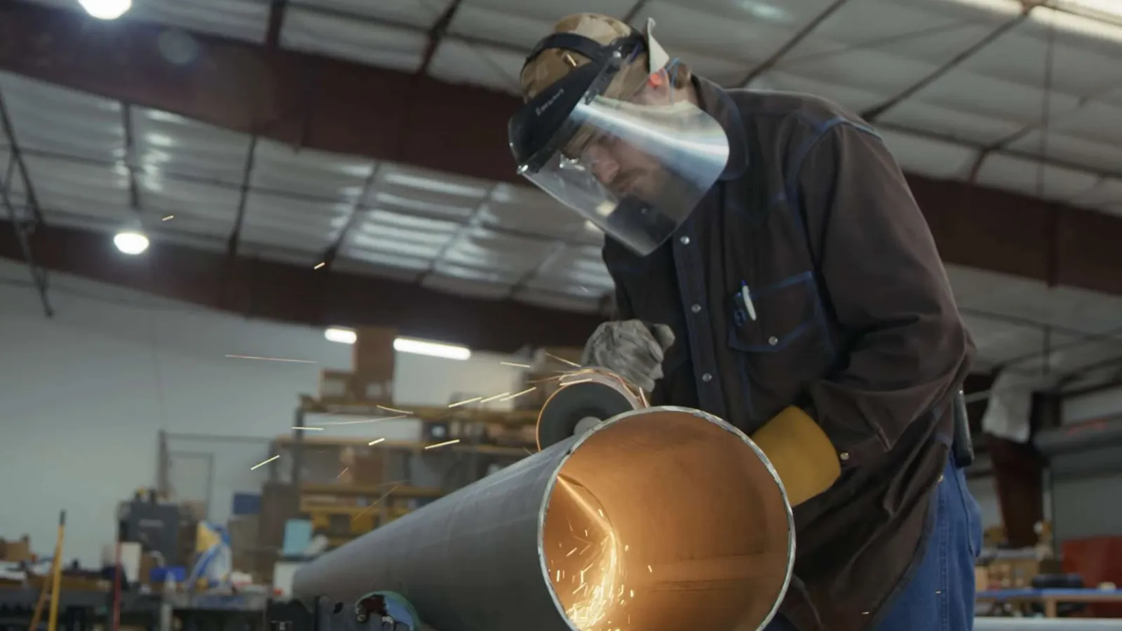 Worker wearing protective gear grinding a large metal pipe with sparks flying in an industrial workshop.