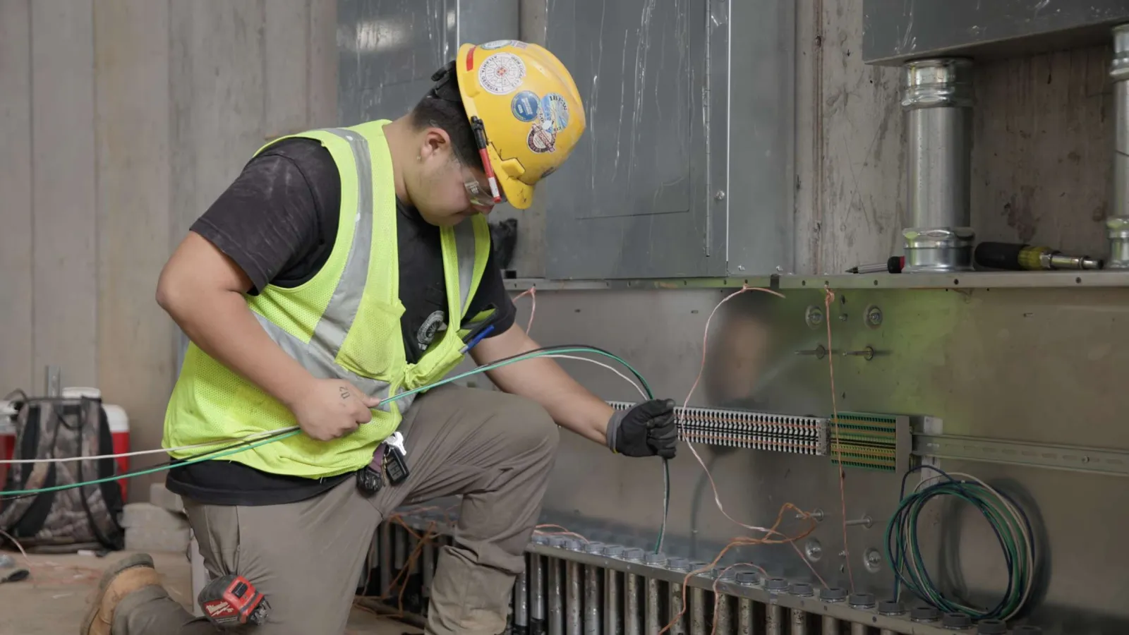 Electrician in safety gear working with wiring inside an industrial electrical panel at a construction site.
