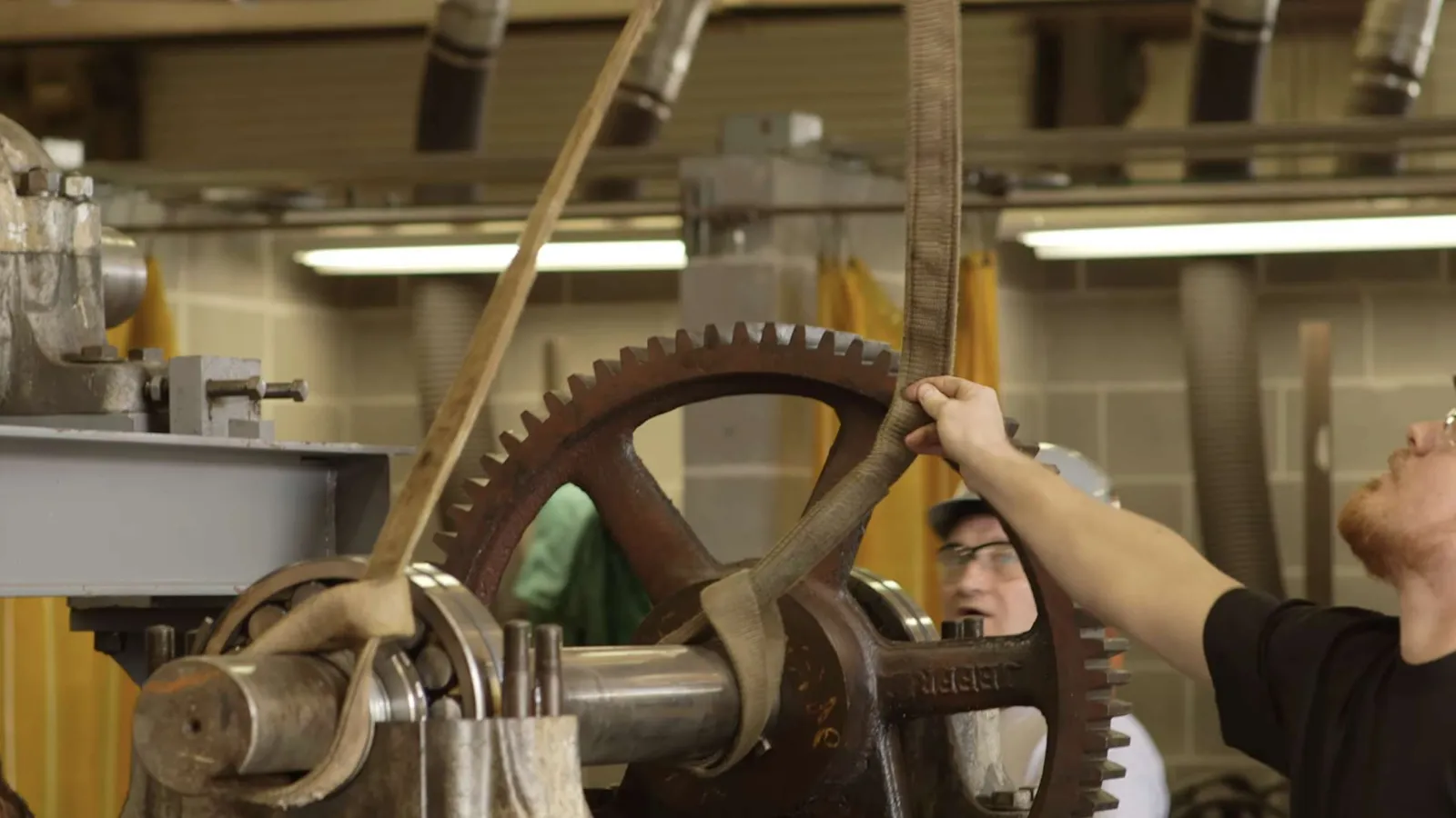 Workers operating and inspecting a large industrial gear mechanism in a factory setting.
