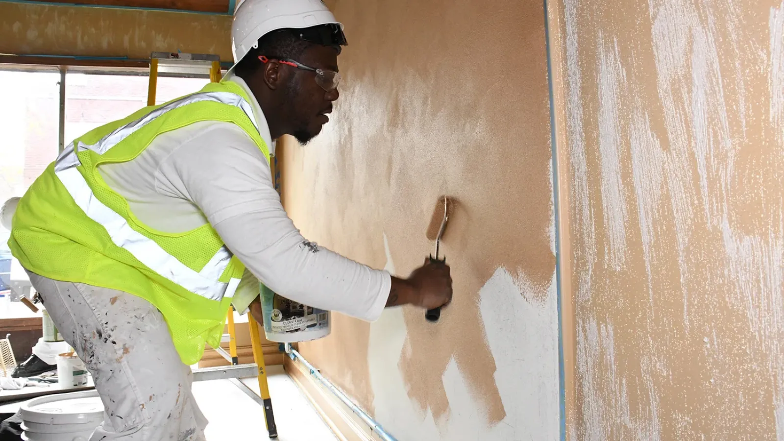 Painter in safety gear applying beige paint to an interior wall with a roller in a construction site.