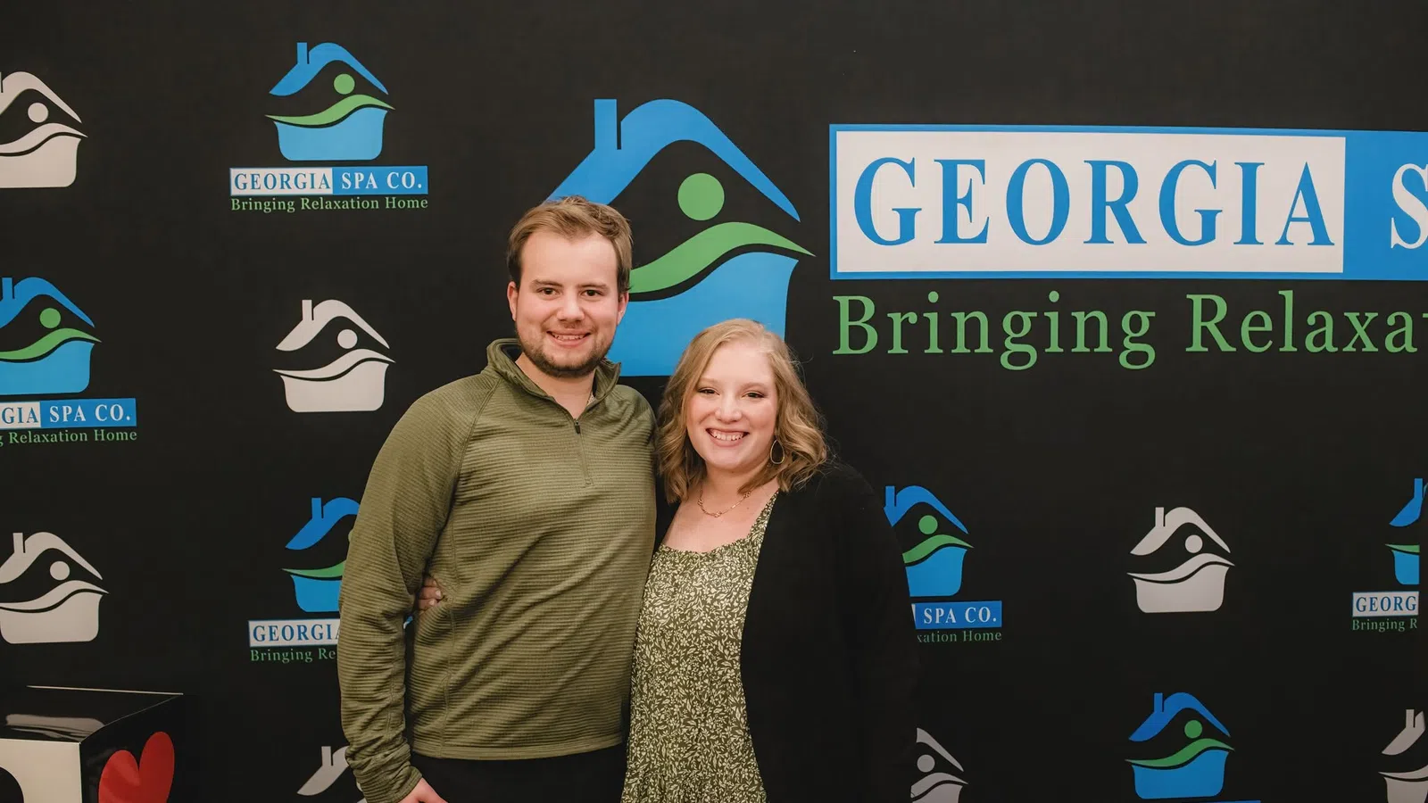 Smiling couple posing in front of Georgia Spa Co. backdrop with logos and relaxation-themed graphics.