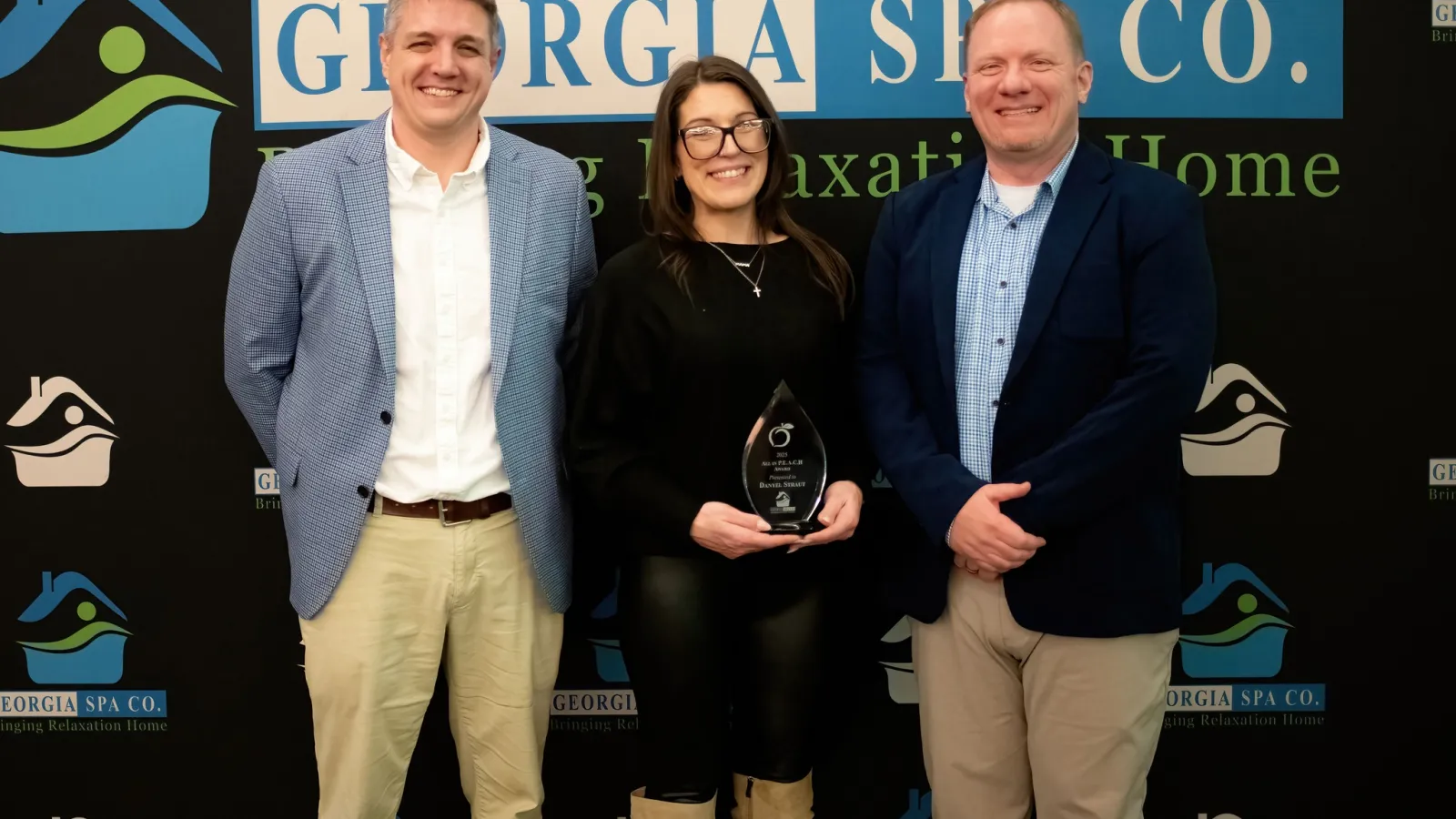 Three people stand smiling in front of a Georgia Spa Co. backdrop with an award held by the woman in the center.