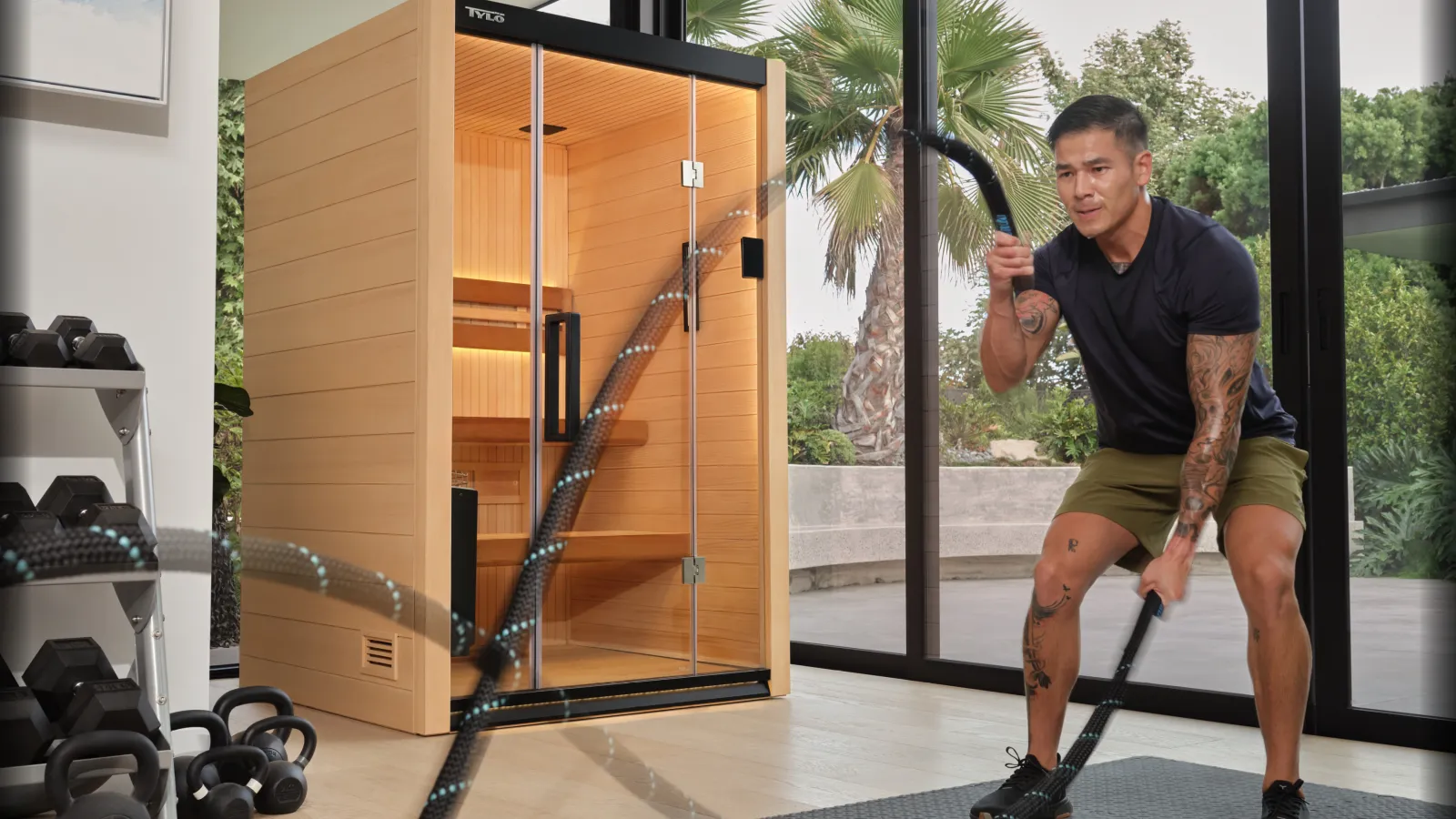 Man exercising with battle ropes in a modern home gym next to a wooden sauna and large glass windows.