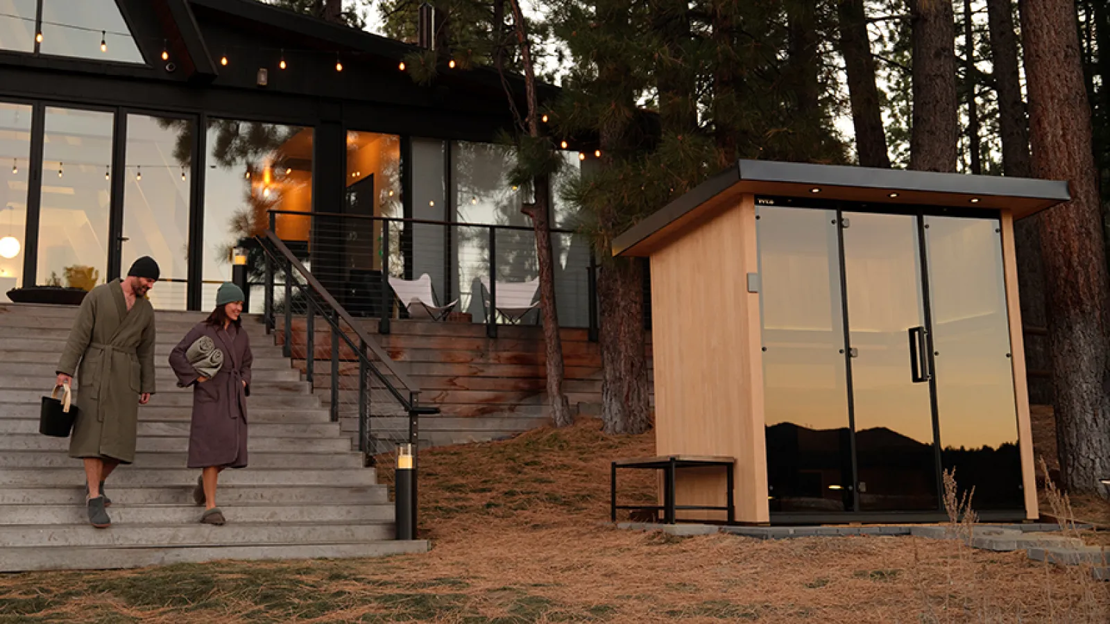Two people walking down steps of a modern cabin in a wooded area at sunset with a small shed nearby.
