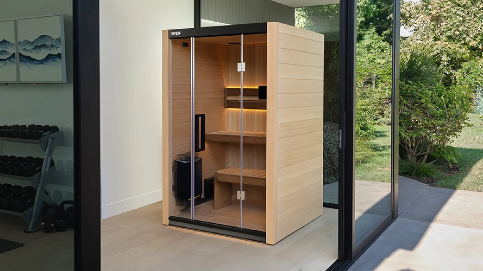 Modern wooden sauna cabin with glass door inside a bright room opening to a green outdoor garden.
