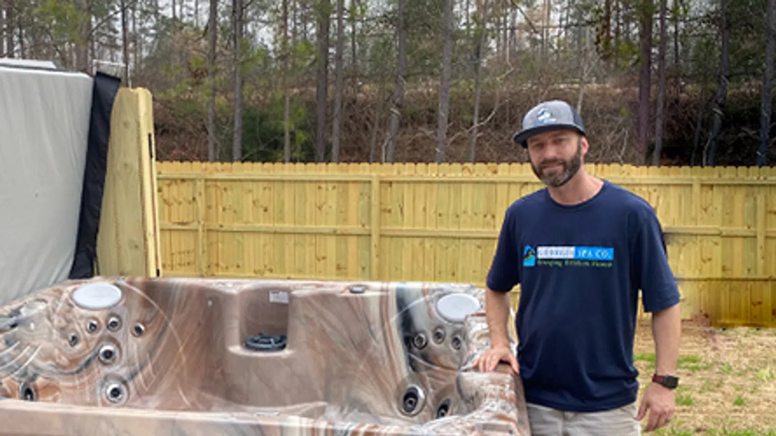Man standing next to a large brown and beige marbled hot tub outdoors by a wooden fence on a concrete patio.
