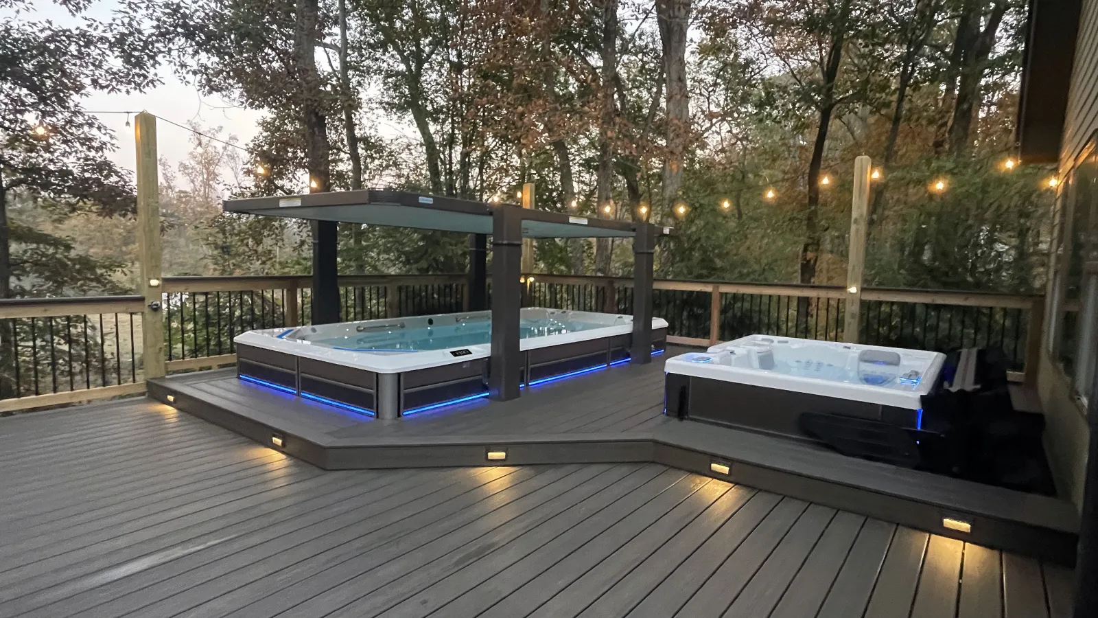 Modern backyard deck with two illuminated hot tubs surrounded by trees and string lights at dusk.