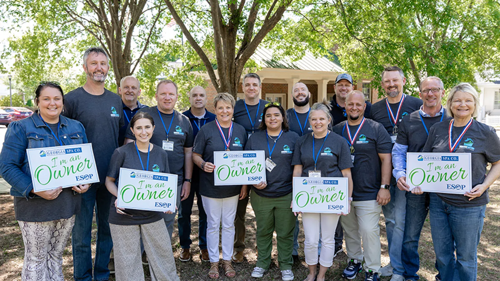 Group of diverse employees holding signs that say 'I'm an Owner ESOP' outside under trees at Georgia Spa Co.