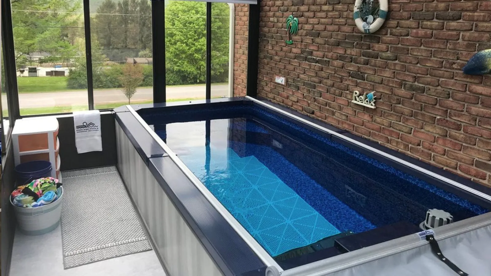 Indoor rectangular swim spa with blue mosaic interior, brick wall decor, and large window view of greenery.