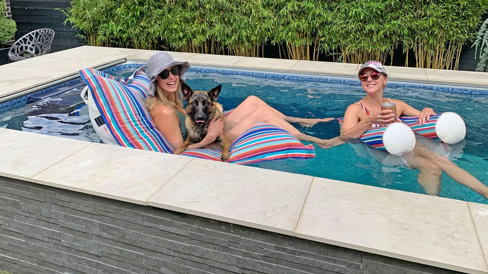 Two women relaxing in a small pool on colorful bean bag floats, one holding a dog, surrounded by bamboo plants.