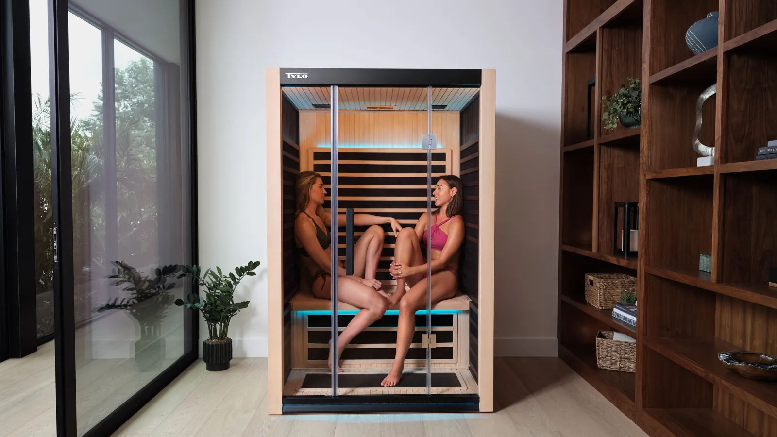 Two women sitting and relaxing inside a modern wooden infrared sauna cabin in a bright room.