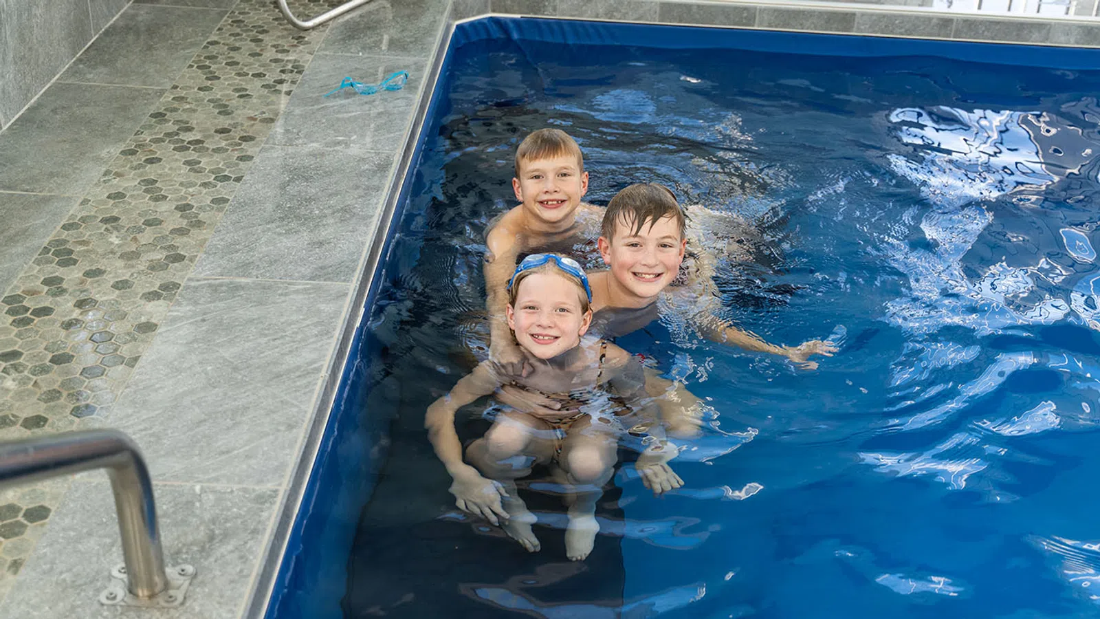 Three smiling children swimming and playing together in a blue indoor pool near tiled edge and metal handrail